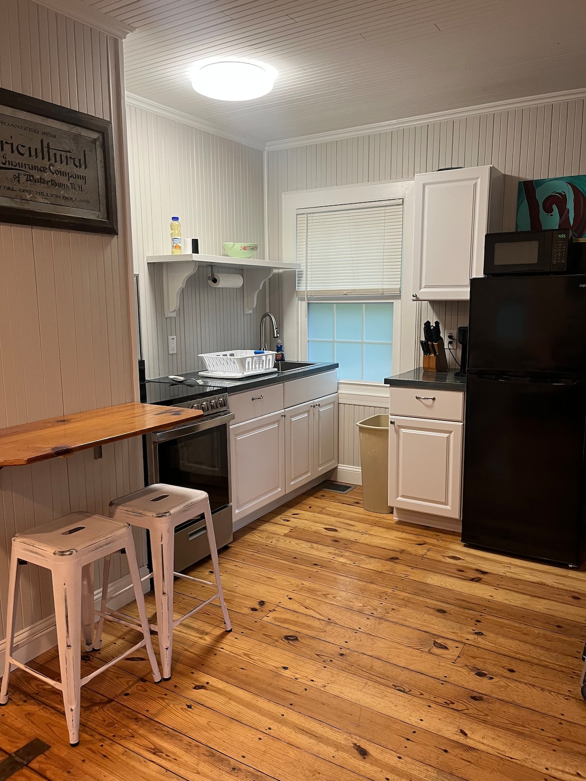 A functional kitchen space is presented with white cabinets and a black countertop. An electric stove and refrigerator are visible, along with an organized sink area. Two metal stools are positioned against a wooden countertop, enhancing the efficient layout of the room.