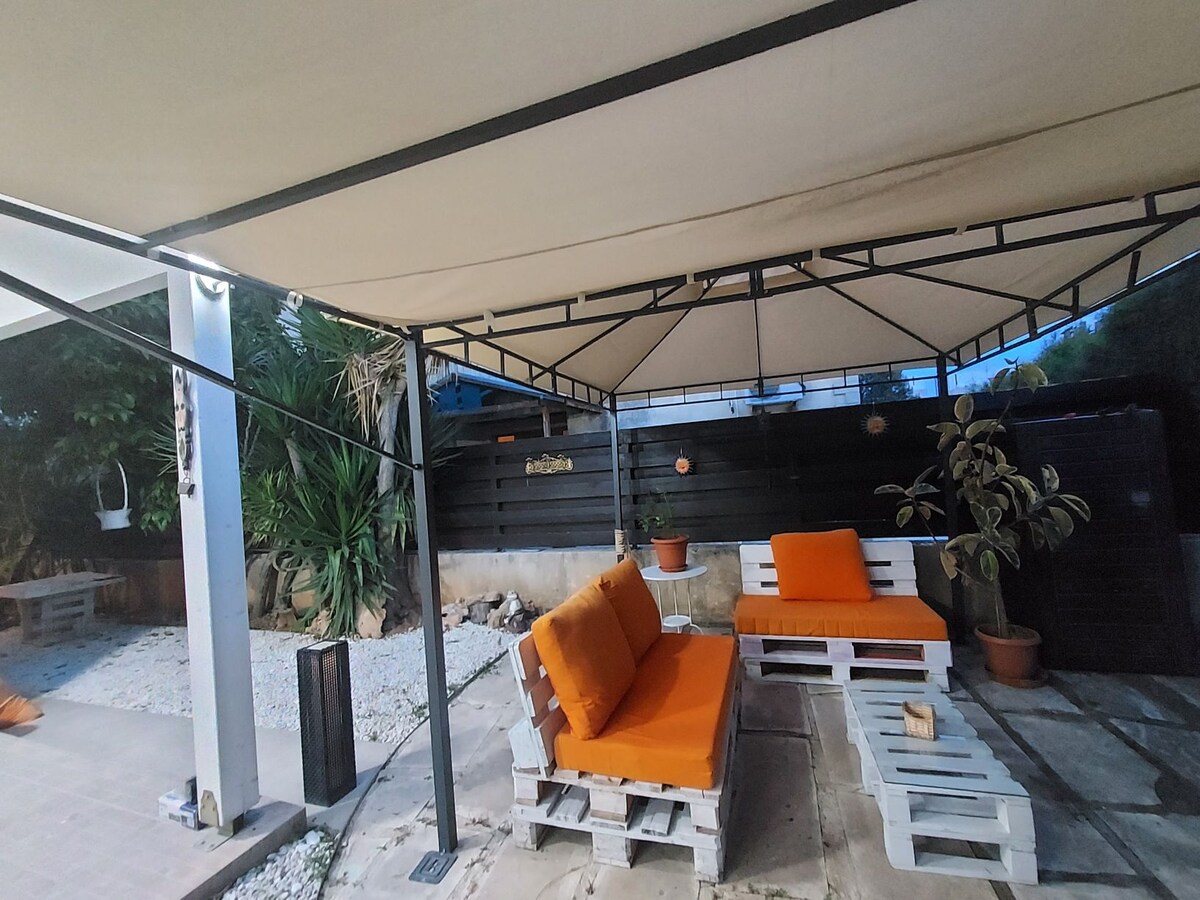 An outdoor seating area features a covered structure with a light-colored canopy. Two orange cushions rest on white wooden seating, complemented by a nearby potted plant. Gravel and concrete provide a minimalist floor, with additional seating visible in the background.