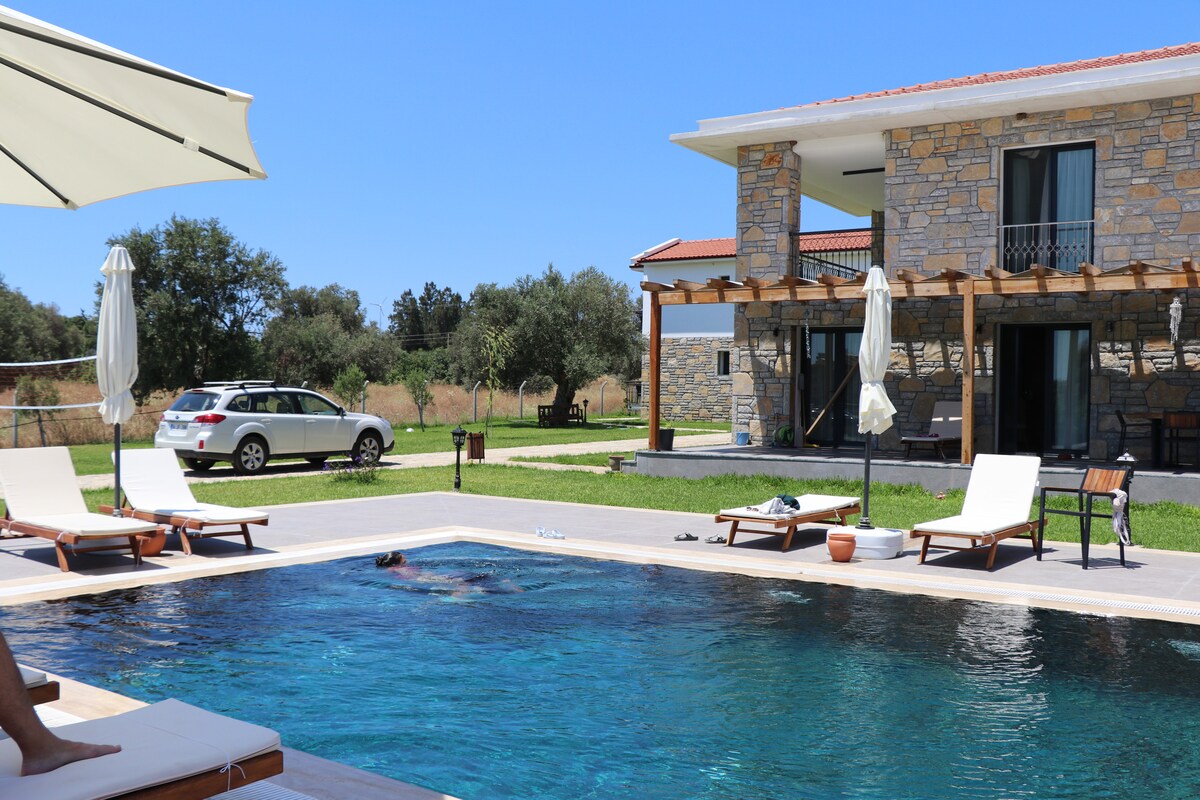 A refreshing outdoor pool is framed by comfortable lounge chairs and umbrellas. A stone house can be seen in the background, surrounded by green grass and trees. A vehicle is parked nearby, adding to the setting's convenience.