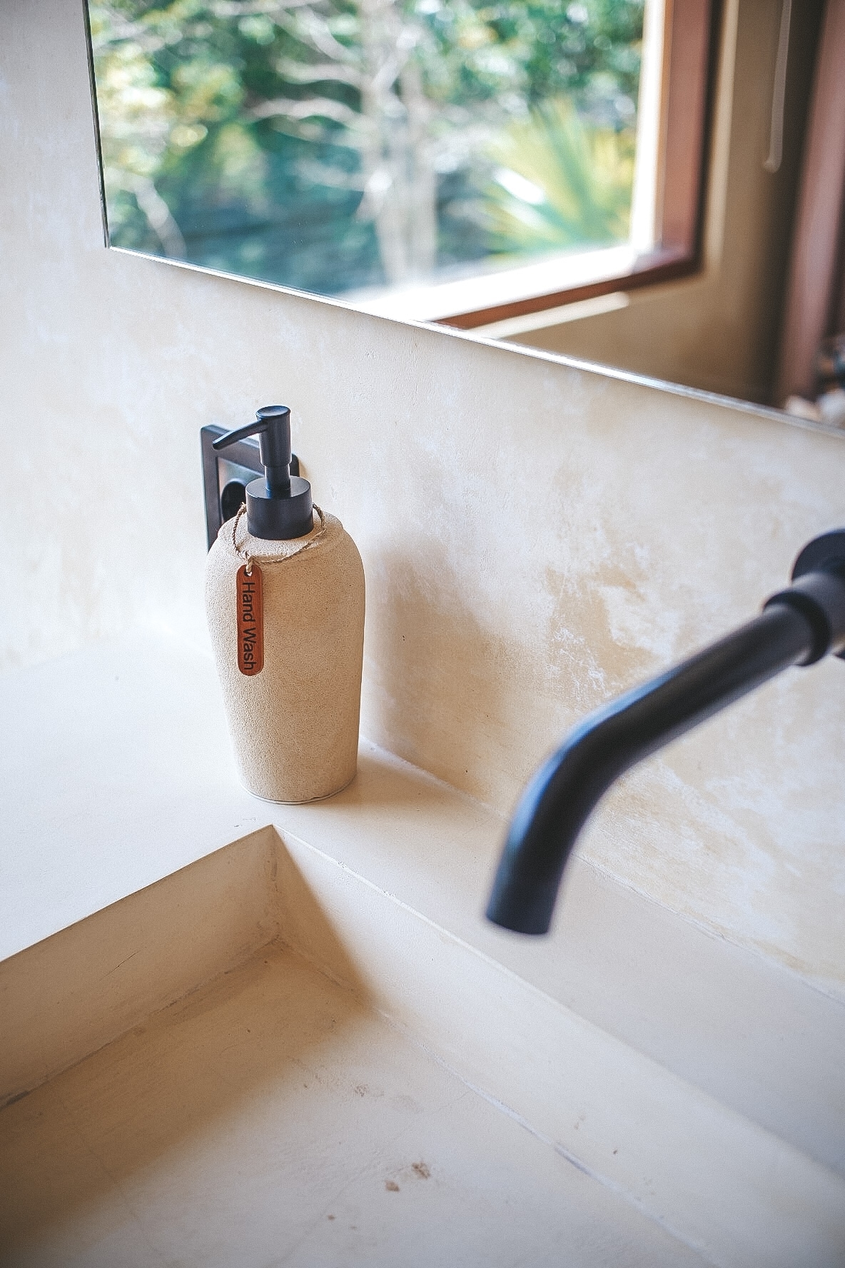 A minimalist bathroom counter is displayed, featuring a stone-like soap dispenser alongside a sleek, matte black faucet. Natural light enters through a window in the background, highlighting the earthy tones of the countertop.