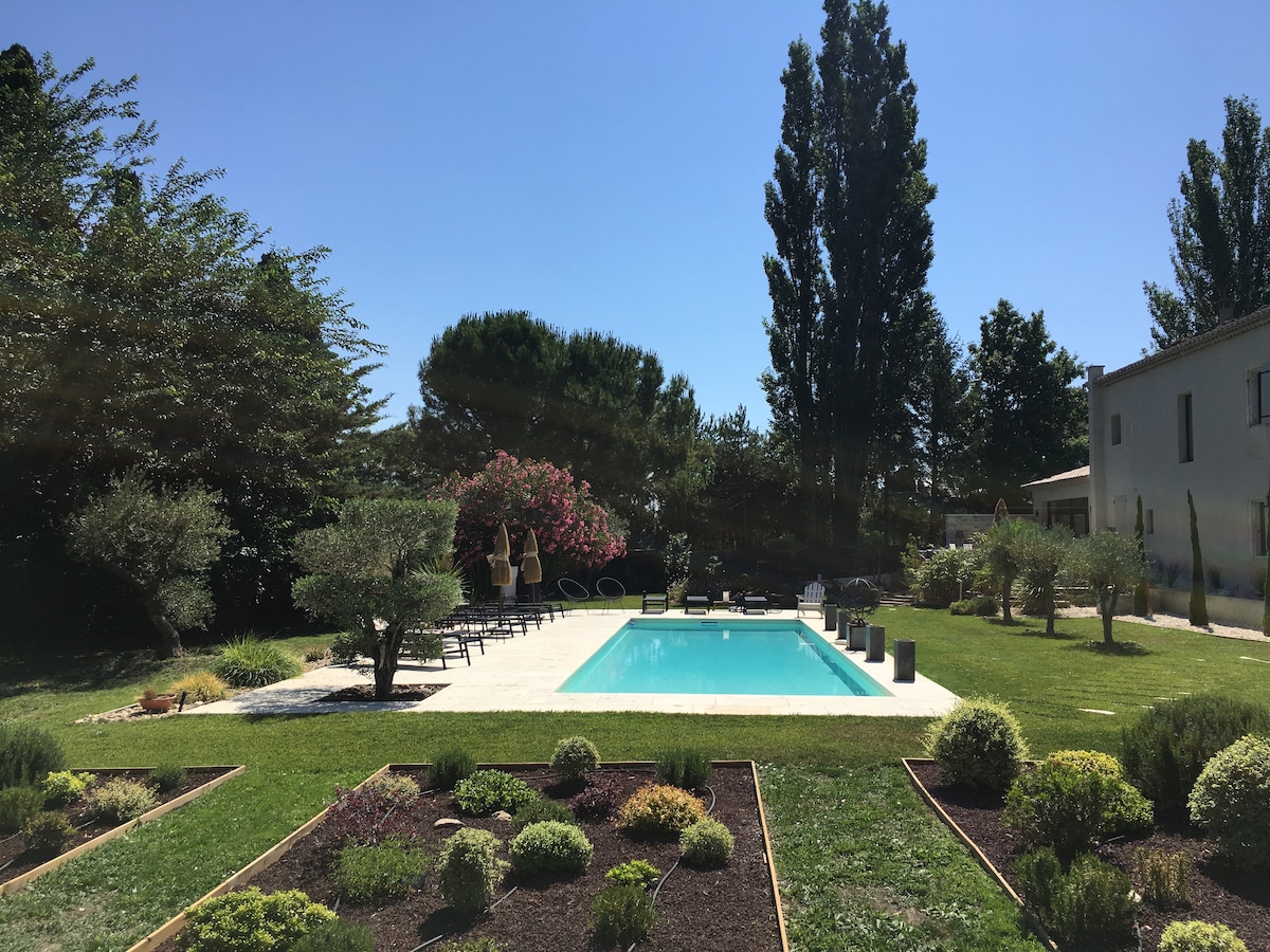 A spacious garden features a rectangular pool surrounded by lounge chairs and shaded areas. Lush greenery, including trees and flowering plants, adds natural beauty, while pathways are lined with well-maintained shrubs. The clear blue sky completes the serene outdoor setting.