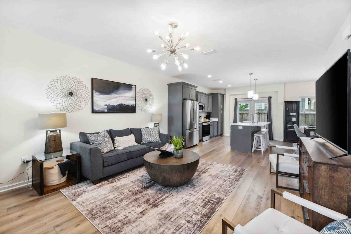 An open-concept living area is presented, featuring a comfortable grey sofa and a central round coffee table. A stylish light fixture hangs above, and a large window allows natural light to fill the space. The modern kitchen, equipped with stainless steel appliances, is visible in the background.