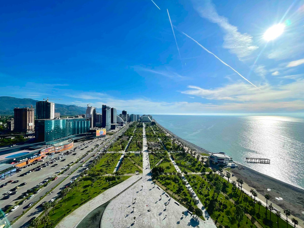 A panoramic view showcases a bustling promenade alongside the sparkling sea. Lush green spaces are interspersed with pathways, leading to the beach. Modern buildings rise in the background under a clear blue sky, while the sun reflects off the water, creating a vibrant atmosphere.