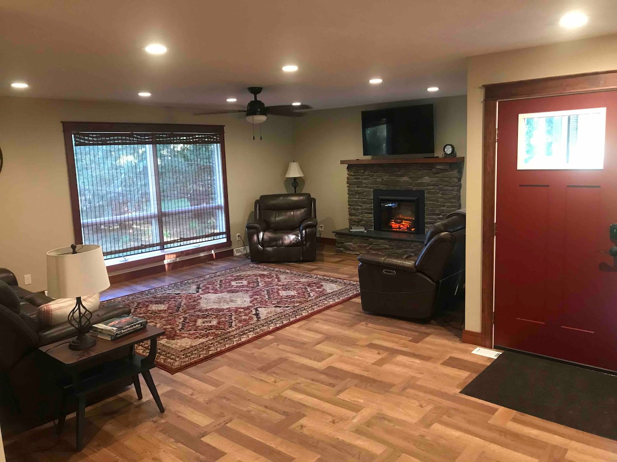 The living room features leather couches arranged around a stone fireplace, with an electric fire providing warmth. Natural light enters through the large windows, partially covered with blinds. A patterned area rug adds a touch of color to the hardwood floor.
