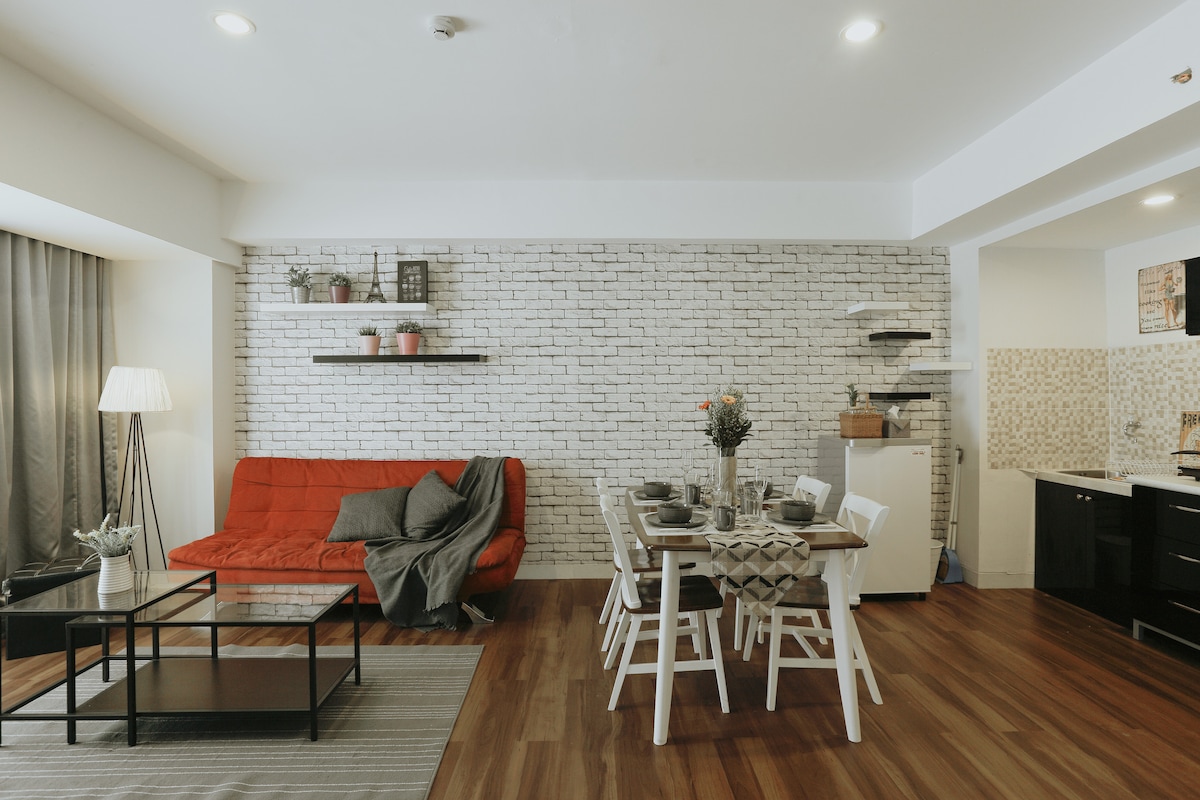 The open living area features a red sofa bed paired with a complementing grey throw. A dining table is set for six, surrounded by white chairs. Shelving on the wall displays potted plants, and a small kitchenette is visible in the background with a refrigerator.
