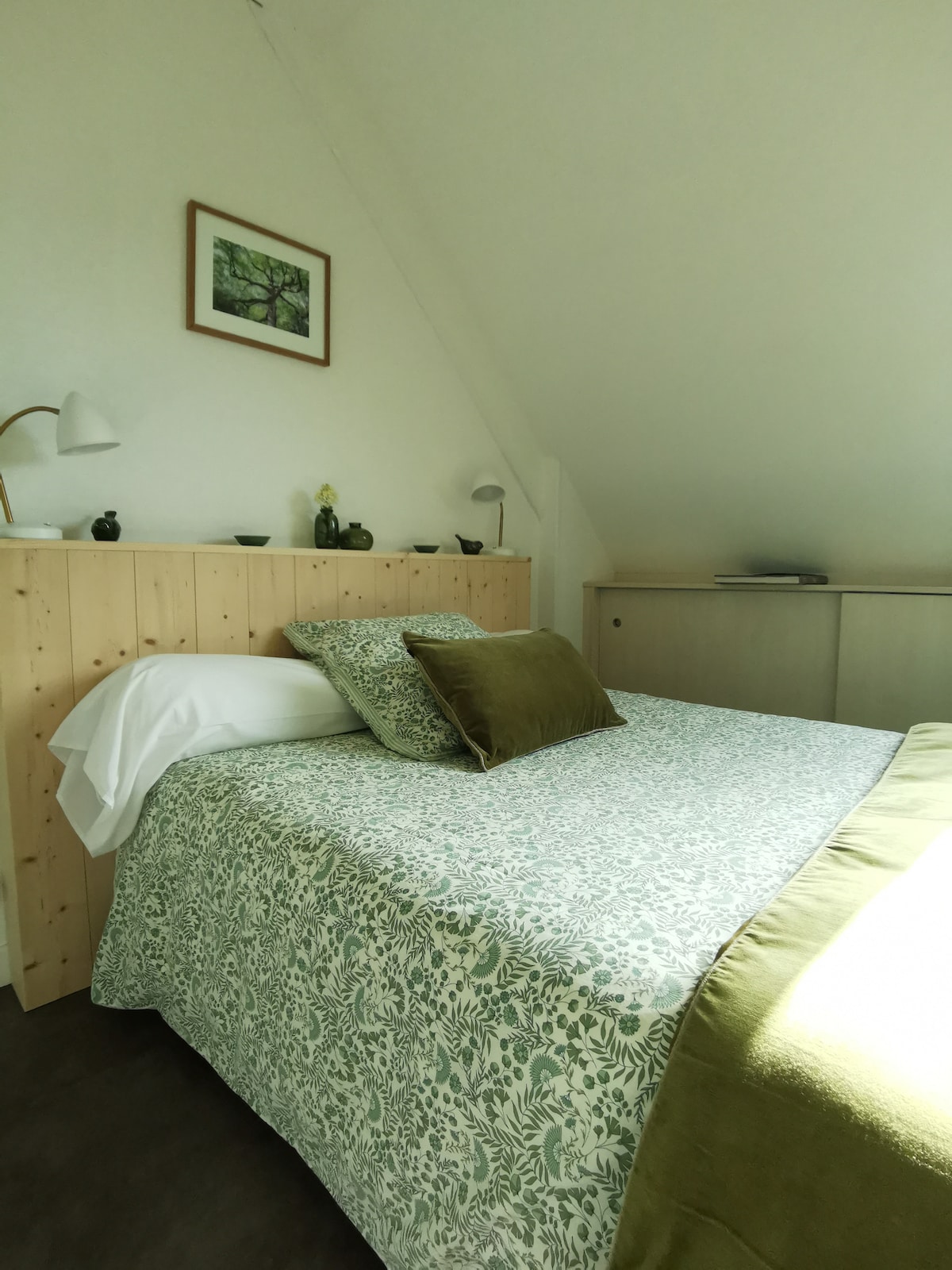 A spacious sleeping area features a light-colored floral bedspread and a decorative green pillow. A wooden headboard adds warmth, while a small bedside lamp and decorative items are placed on the side. A neutral cabinet is visible in the background, contributing to the room's serene ambiance.