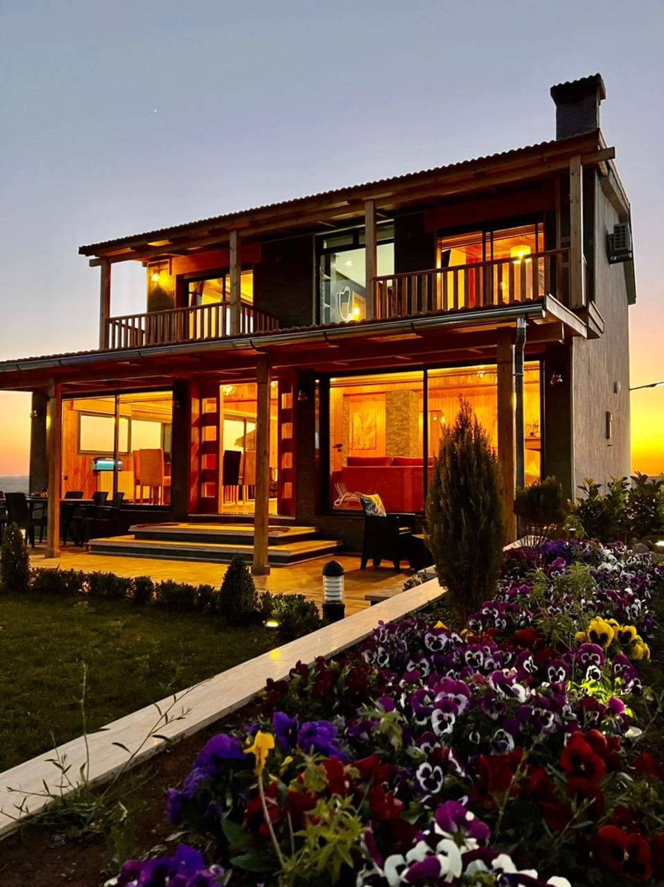 A two-story chalet is showcased at dusk, featuring large glass doors that provide a view of a welcoming interior. A vibrant garden with colorful flowers surrounds the property, and warm light emanates from inside, enhancing the cozy atmosphere of the wooden structure.