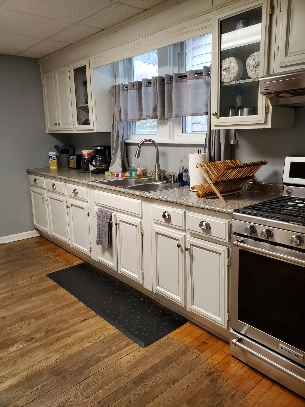 A fully equipped kitchen features white cabinetry and dark countertops. A stainless steel stove is positioned beside a microwave. A coffee maker and dish rack are visible on the countertop, with a window above the sink allowing natural light to enter.