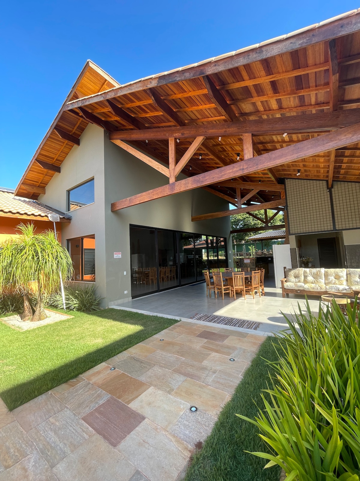 The exterior of the house is characterized by a striking roof design and spacious glass walls, allowing for abundant natural light. A pathway of stone tiles leads to a well-kept lawn, framed by lush greenery, and outdoor seating is visible under the covered patio.