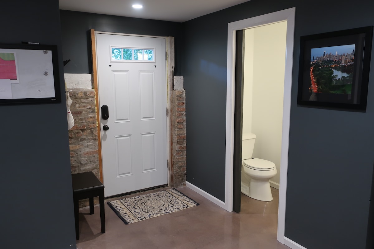 An entryway is presented, featuring a dark blue wall contrasted by a light-colored door with a large window. A small table is positioned to the left, and a rug lies beneath. A bathroom is visible through a doorway, displaying a white toilet against a light background.