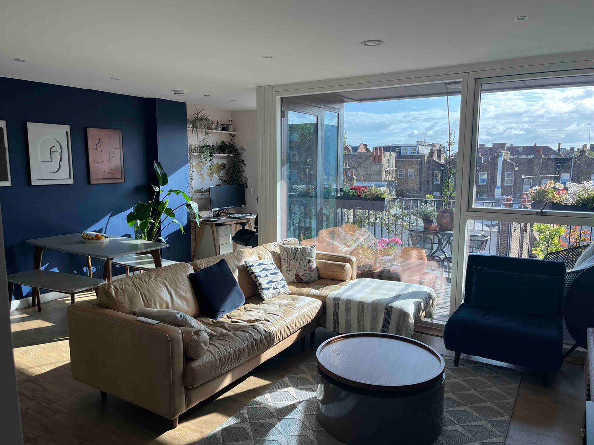 A spacious living area features a light-colored sofa with patterned cushions, complemented by a round coffee table. A blue accent wall enhances the contemporary decor. Large windows provide natural light and a view of the balcony with flowers, along with a dining table positioned nearby.