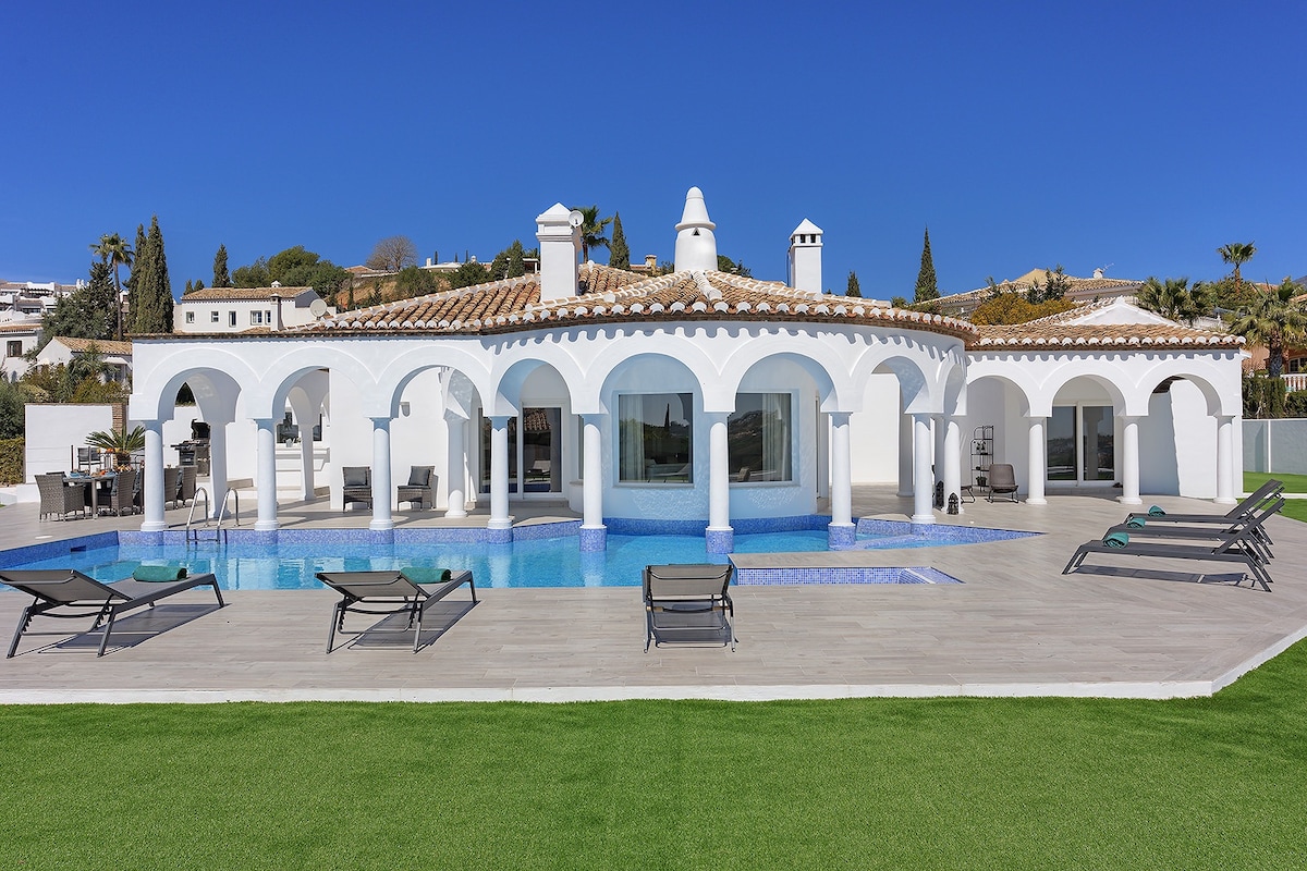 A modern villa features a spacious outdoor area with a large saltwater pool, lounge chairs, and an expansive lawn. The building showcases arched doorways and a bright white exterior, complemented by clear blue skies and surrounding lush landscaping.