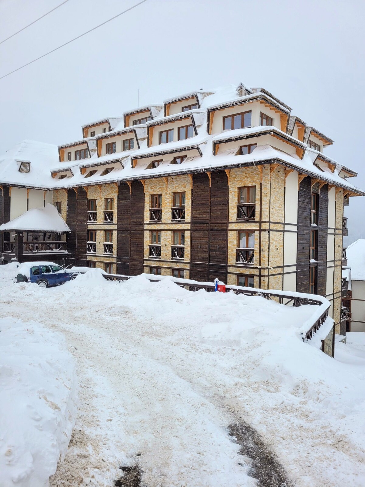 A modern hotel building is visible, characterized by a mix of wood and stone materials. Snow covers the ground, creating a serene winter landscape. The structure features multiple levels, with a distinctive roofline and spacious balconies that provide views of the surrounding area.