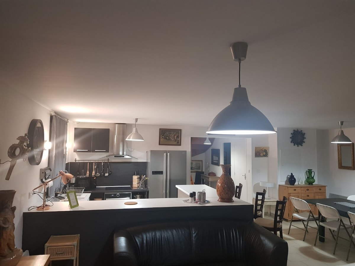 An open-plan kitchen and dining area are highlighted by pendant lighting. A dark leather couch is positioned in the foreground, while the kitchen features modern appliances, including a refrigerator and stove. A wooden dining table with chairs is visible in the background, enhancing the social atmosphere.