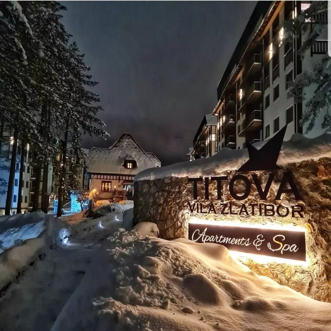 The entrance to Titova Apartments and Spa is shown, with a snow-covered pathway leading up to it. Warm lighting illuminates the sign against a backdrop of snowy trees and a charming building, contributing to a serene winter atmosphere.