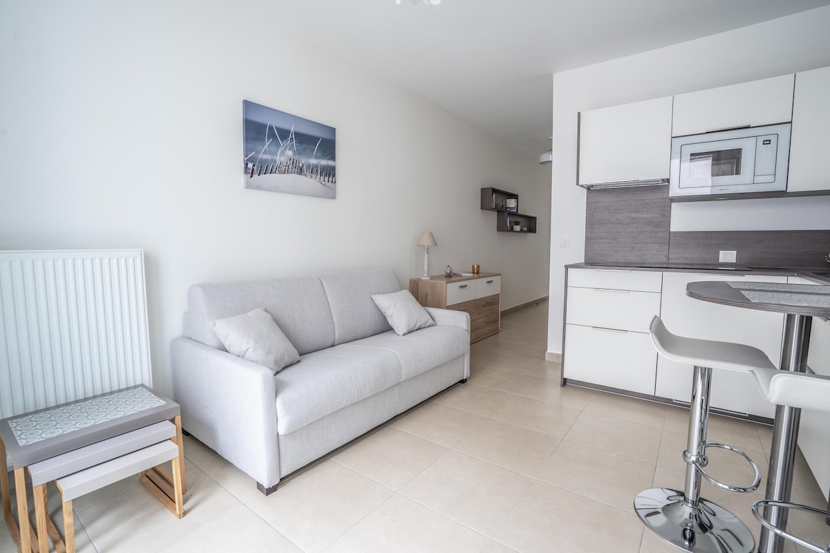 A modern living area is showcased with a light-colored sofa and a minimalistic table. A microwave is built into the cabinetry, and additional storage units are visible. Soft, natural light illuminates the space, complementing the neutral color palette. A decorative piece hangs on the wall.
