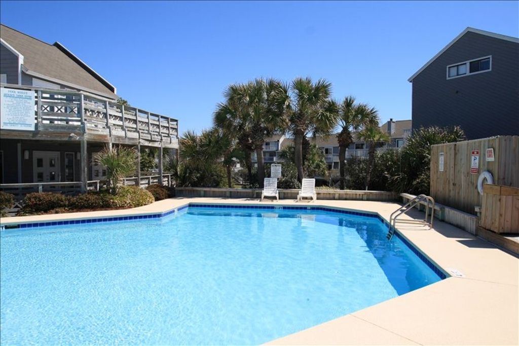 A large outdoor swimming pool is depicted, surrounded by vibrant palm trees and tropical vegetation. Two lounge chairs are positioned near the pool's edge, with a fenced area visible in the background. Clear blue skies and bright sunlight create a refreshing atmosphere.