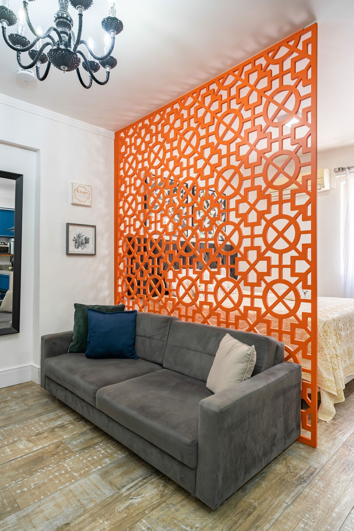 A modern gray sofa is positioned against a vibrant orange decorative panel, which features intricate geometric patterns. A framed artwork is mounted on the wall, and soft lighting is provided by a stylish chandelier above, creating a unique separation between the living area and sleeping space.