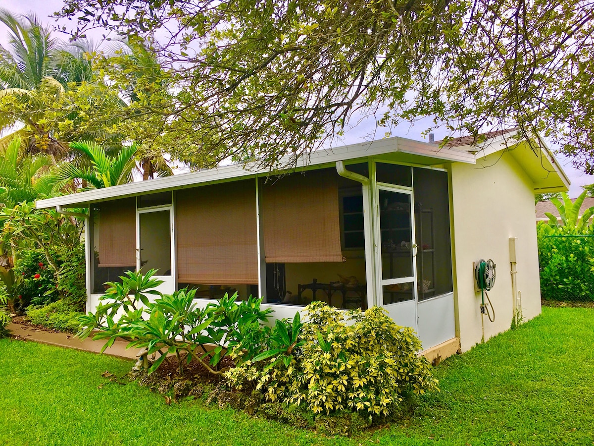 The exterior of a private guesthouse is showcased, surrounded by lush greenery and tropical plants. Screened windows provide a view of the outdoors while offering a sense of openness. The building is designed with a light-colored façade, blending harmoniously with the natural setting.