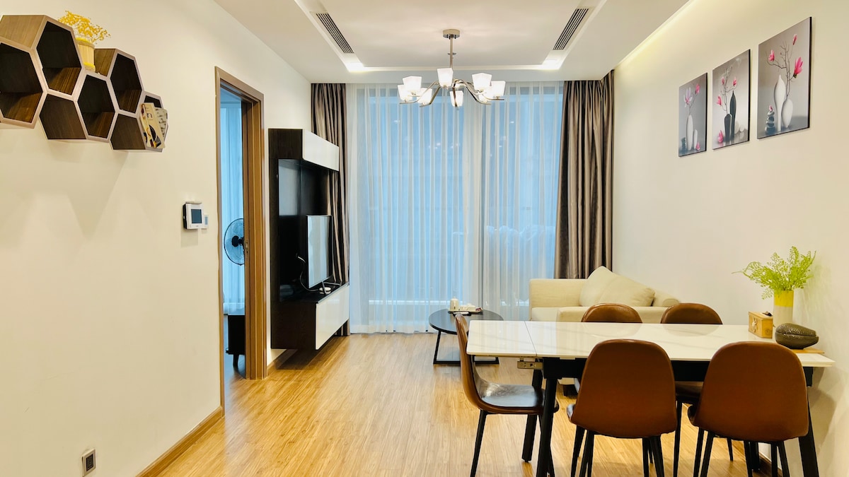 The well-lit living area features a comfortable sofa and a dining table with chairs. A media unit is positioned against the wall, and sheer curtains cover the windows, allowing natural light to filter in. Wall art adds a touch of elegance to the space.
