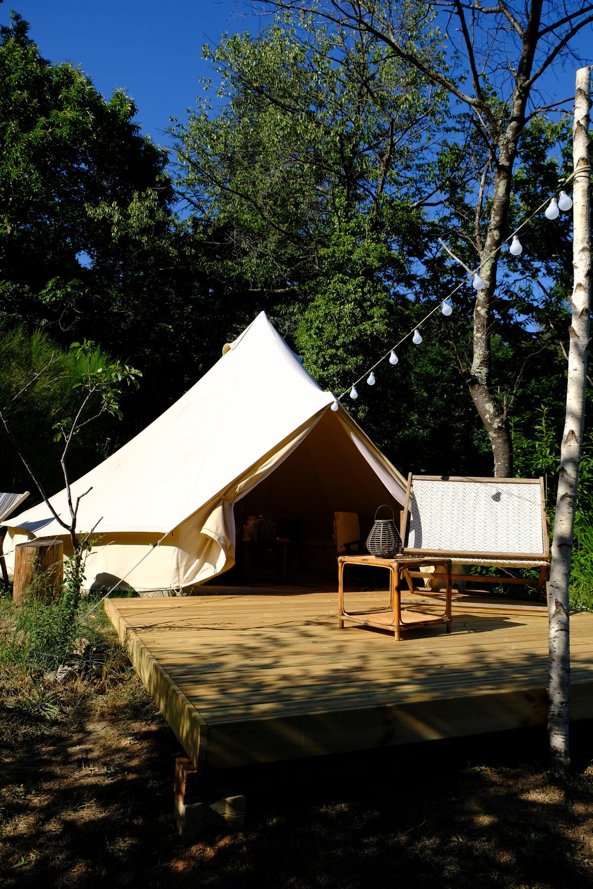 A spacious tent is set on a wooden platform, surrounded by greenery. A small wooden table and chair are positioned outside the tent entrance, while decorative lights hang above the area, creating an inviting outdoor space.
