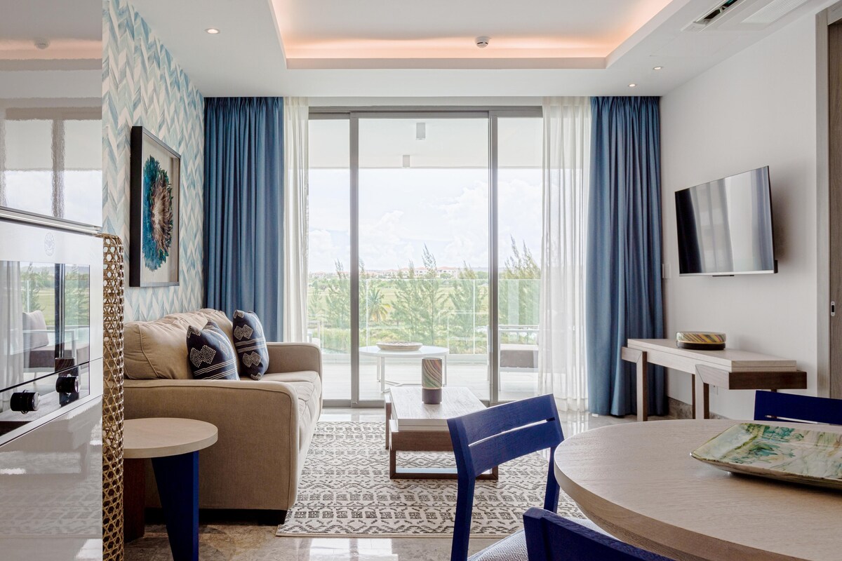 A modern living area features a neutral-toned sofa, accented by colorful throw pillows. Large glass doors open to a balcony, allowing natural light to fill the space. A sleek television is mounted on the wall, while a dining table is complemented by blue chairs.