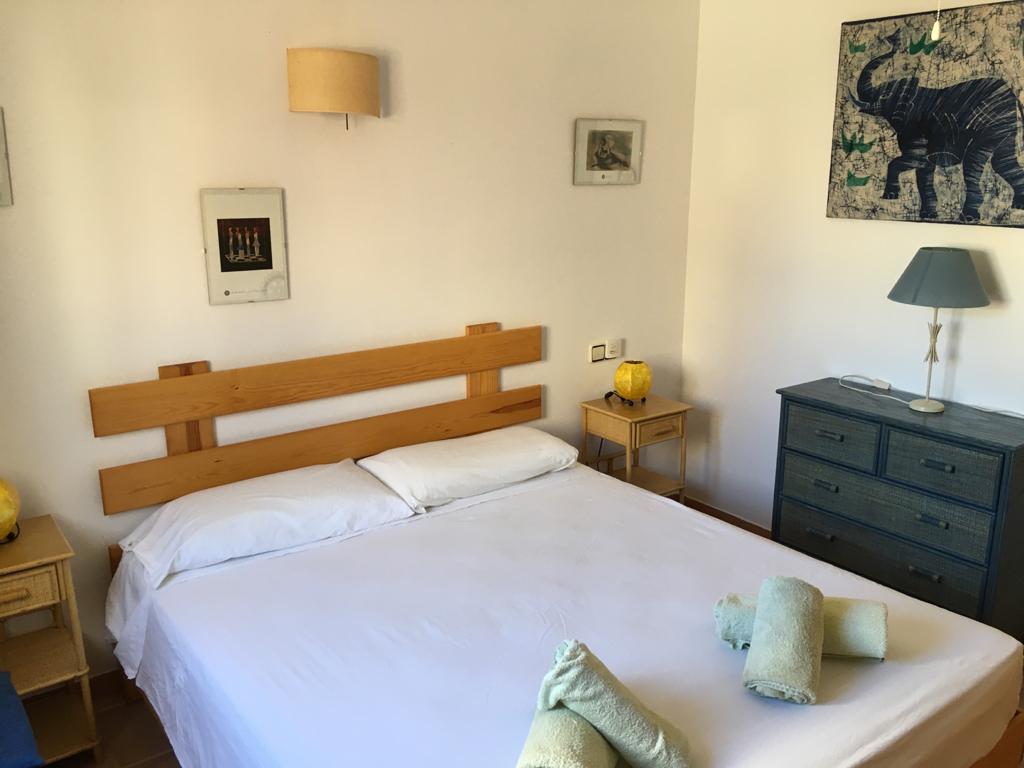A simple bedroom is furnished with a spacious bed dressed in white linens. Two rolled towels are neatly placed at the foot of the bed. A blue lamp and artwork adorn the bedside tables, while a dark chest of drawers offers additional storage beside a framed image.