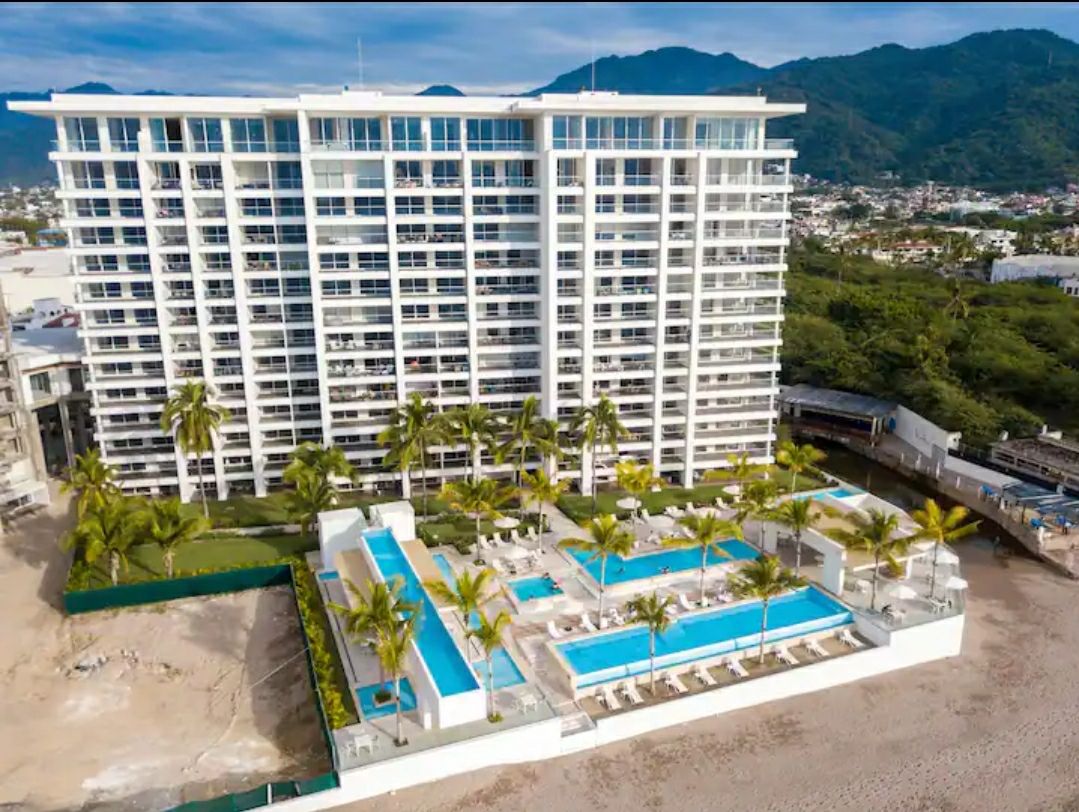 An aerial view of a modern multi-story building is visible, surrounded by lush palm trees. Multi-level pools are showcased in the foreground, featuring clean blue water and numerous lounge chairs arranged along the edges, creating an open and inviting leisure area.