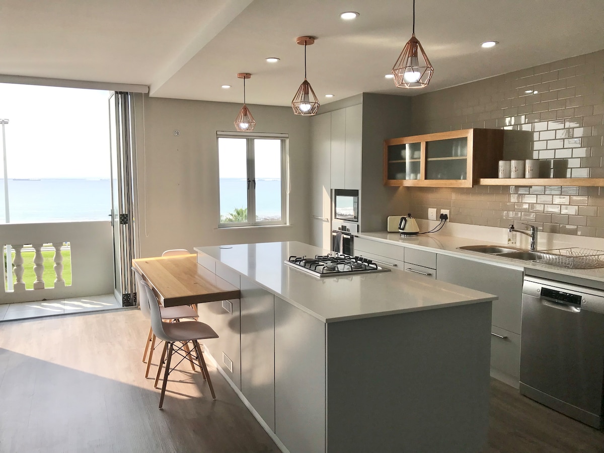 A modern kitchen is displayed with sleek cabinetry and a spacious island. Three pendant lights are hung above the island, providing illumination. A dining table with two chairs is positioned nearby, and large windows offer views of the sea and beach.