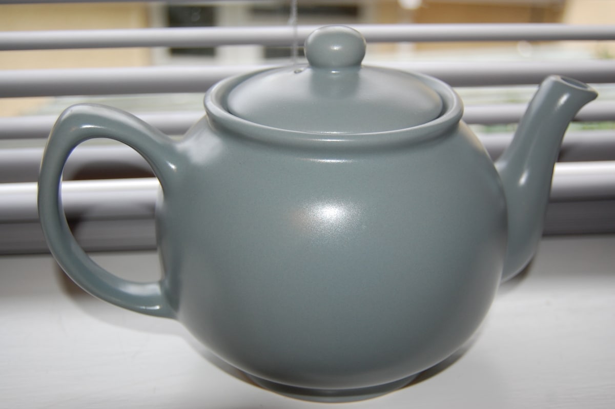 A gray ceramic teapot is displayed on a windowsill, featuring a rounded body and a curved spout. The lid is securely positioned, and the handle is seamlessly integrated. Light filters through the closed blinds, creating a soft atmosphere around the teapot.