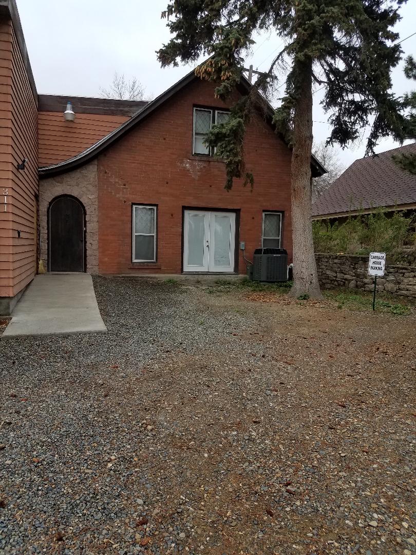 The exterior of the house features a combination of brick and wood siding, with a large, arched entryway. A gravel pathway leads to the entrance, surrounded by greenery. An air conditioning unit is positioned beside the house, and off-street parking is available.