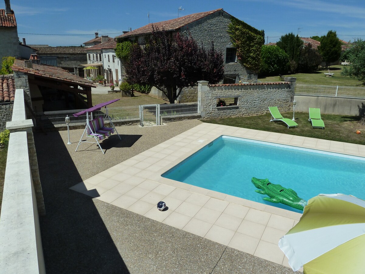 A shared pool area features a rectangular pool surrounded by light-colored tiles. Lounge chairs are positioned nearby, complemented by a beach umbrella. Lush greenery and rustic buildings are visible in the background, enhancing the peaceful atmosphere of the outdoor space.