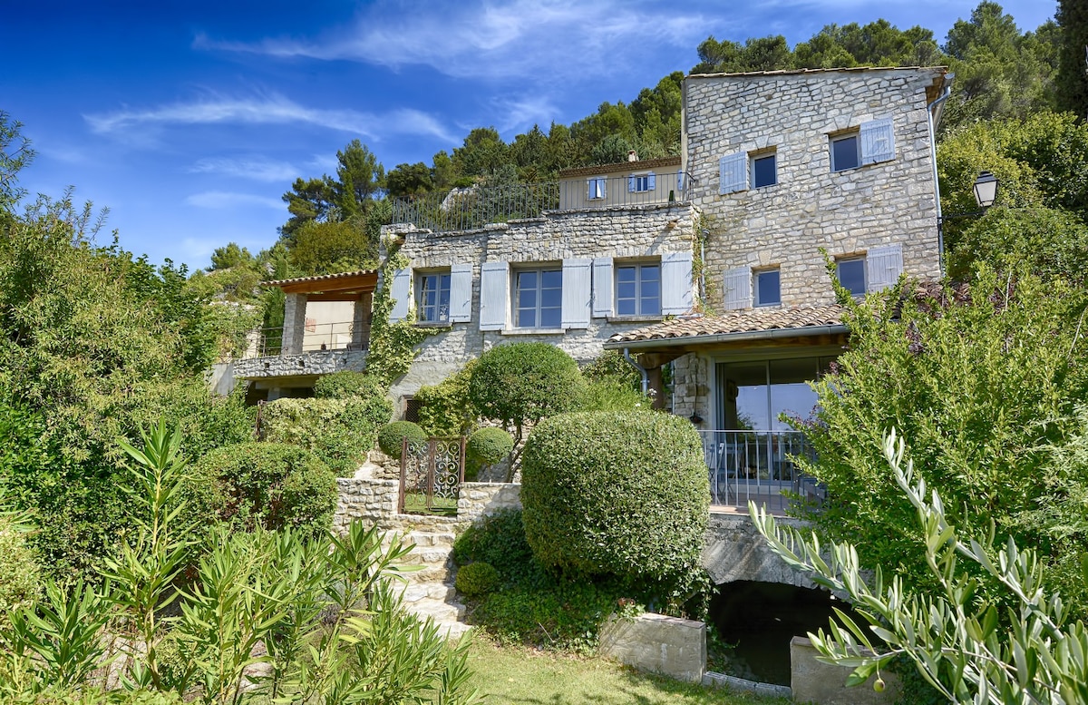 The stone house exhibits a blend of rustic and elegant architecture, surrounded by well-maintained gardens. Lush greenery frames the entrance, while multiple windows enhance the natural light. A terrace can be seen above the garden area, offering a space for relaxation and outdoor enjoyment.