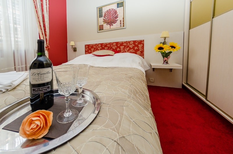 The bedroom features a spacious bed with a decorative headboard, complementing the red accent wall. A tray with two glasses, a bottle of wine, and a rose is placed on the bed, while a vase of sunflowers adds a touch of brightness to the room.