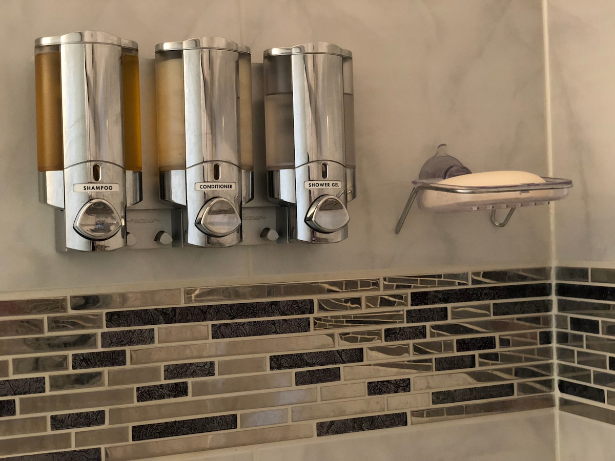 A shower wall displays three chrome dispensers labeled for shampoo, conditioner, and shower gel, positioned beside a clear shelf with a bar of soap. The background features a mix of gray and silver tiles, enhancing a clean and modern appearance.