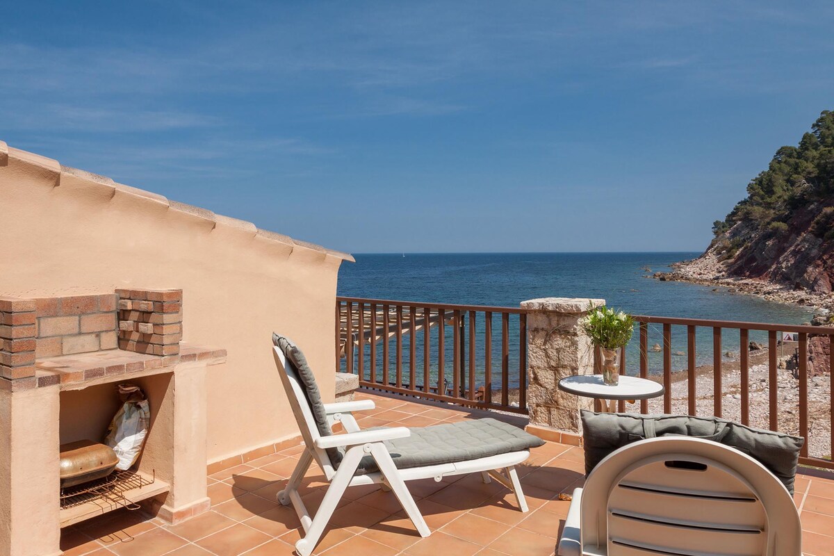An outdoor terrace features a sun lounger and a small table set against a backdrop of the sea. Nearby, a barbecue area is visible, and natural stone cliffs rise in the distance. Clear blue skies create a serene coastal setting.