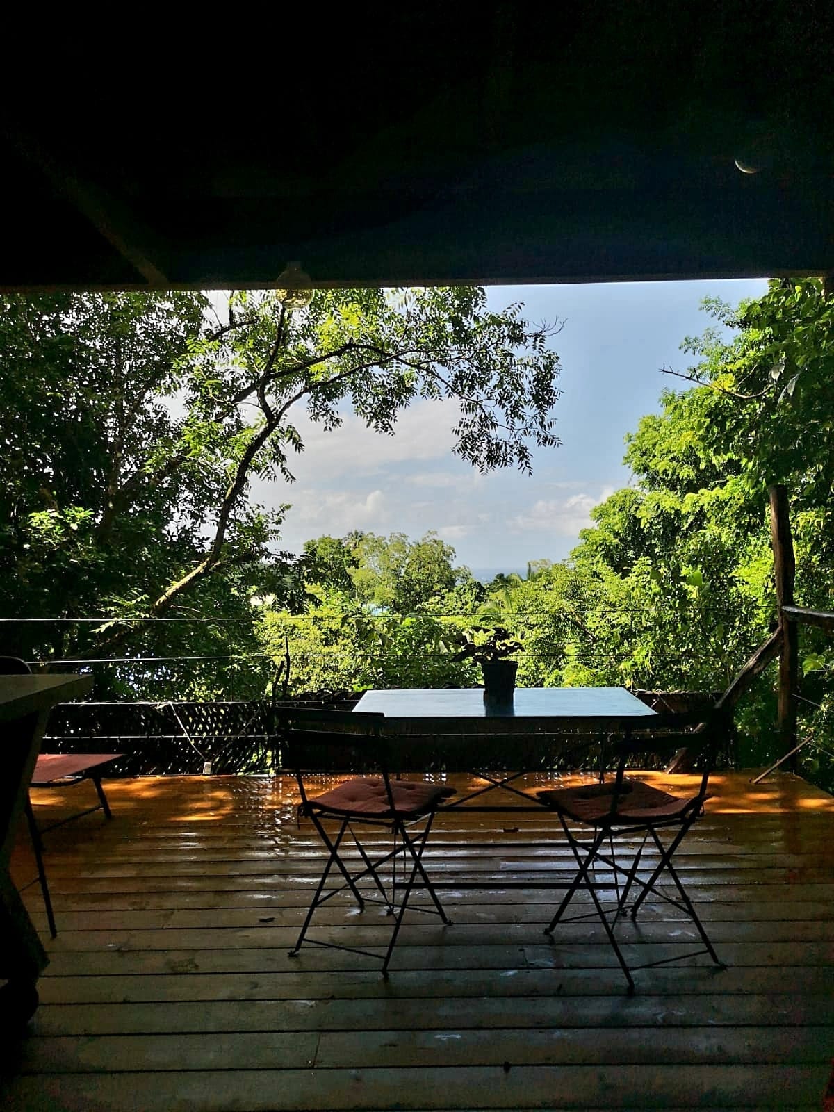 A cozy terrace is viewed, surrounded by lush greenery. A small table with two chairs is positioned on a wooden floor, creating an inviting space for relaxation. The horizon displays a glimpse of the sea under a bright sky, framed by the natural landscape.