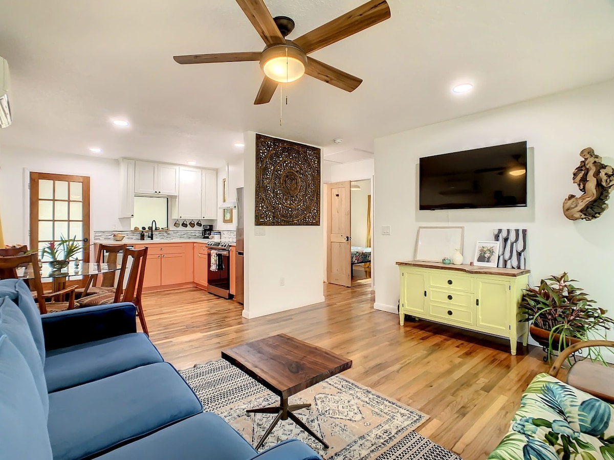 The living area features a blue sectional sofa paired with a wooden coffee table, set on a patterned rug. A ceiling fan hangs above, while a television is mounted on the wall. The open layout connects to a kitchen area with modern appliances and a warm, inviting decor.
