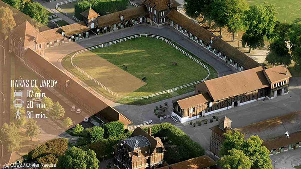 An aerial view of a horse riding facility is displayed, featuring an oval arena surrounded by lush greenery. Several stable buildings are visible, with a neatly maintained layout. The area appears inviting for equestrian activities, with clear pathways and fencing enhancing the organized appearance.