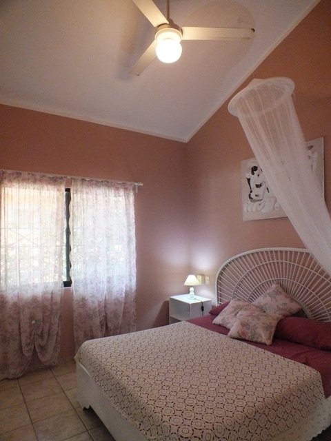 A softly lit bedroom features a spacious bed with a patterned cover and floral pillows. Light pink walls enhance the warm atmosphere. Sheer curtains frame a window, with a bedside table and lamp positioned nearby. A ceiling fan is mounted above, providing comfort.