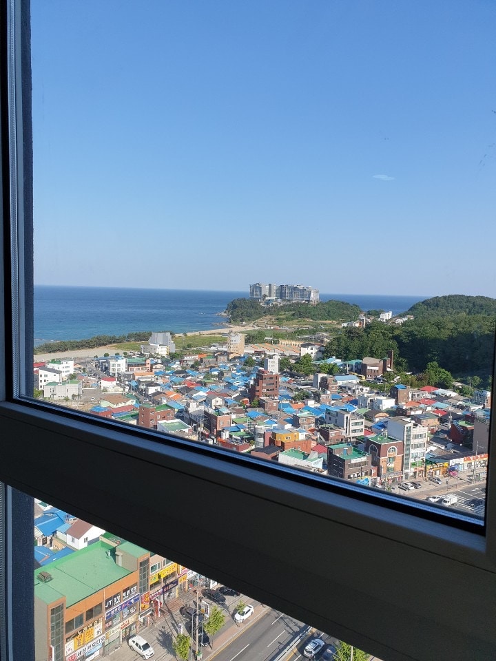 A panoramic view from a window showcases a vibrant coastal landscape. Colorful buildings dot the foreground, leading to a serene stretch of beach and the ocean beyond. A distant hillside features urban structures, framed by clear blue skies.