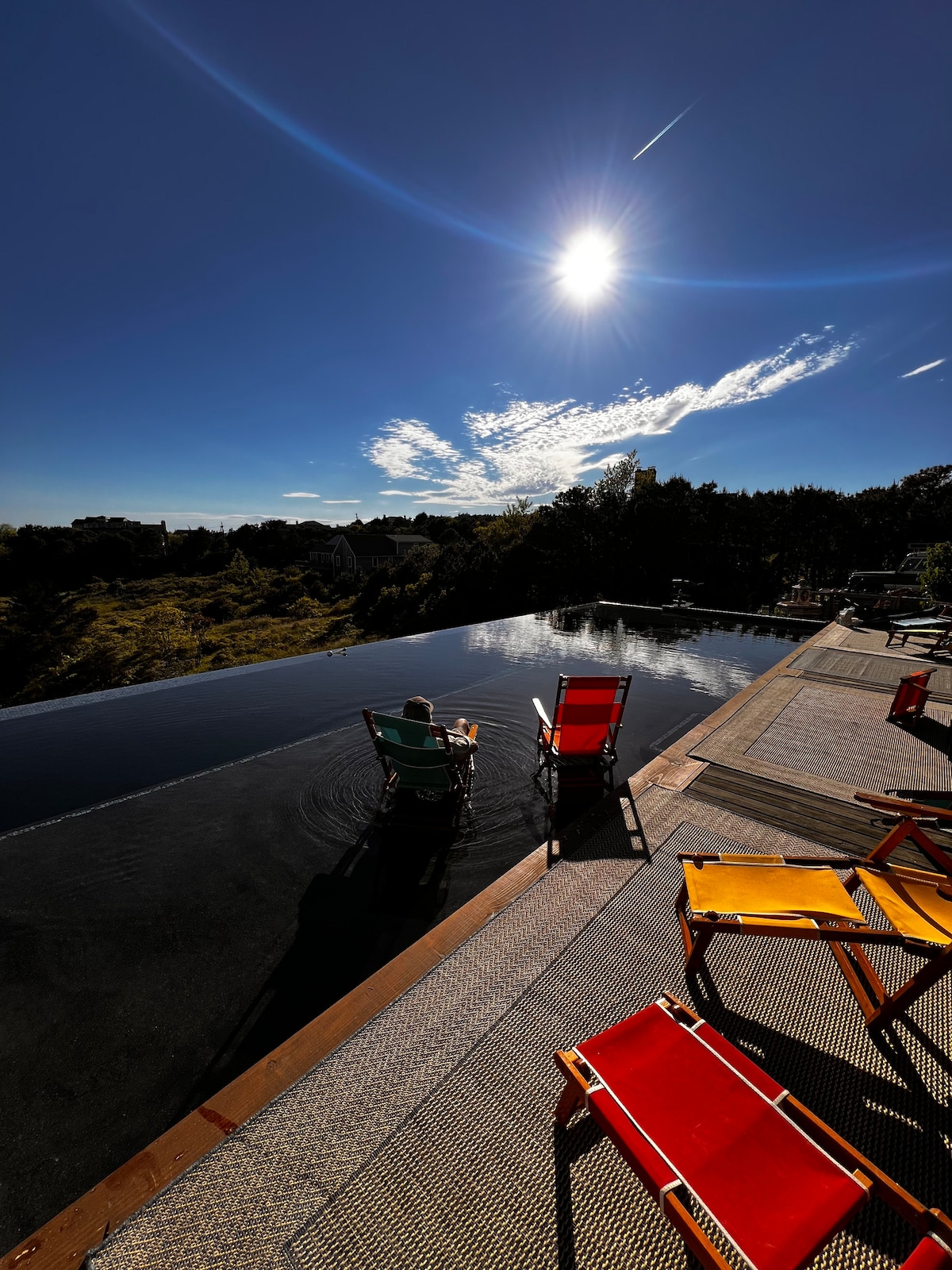 Two colorful lounge chairs are positioned by the edge of a heated pool, reflecting the clear blue sky and vibrant sunlight. The pool's surface mirrors the surrounding lush landscape, while a figure is seen sitting in one chair, enjoying the serene atmosphere.