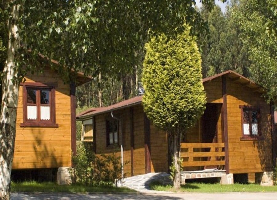 Two wooden bungalows are nestled among lush green trees, featuring distinct facades with brown window frames and porches. A neatly paved walkway leads to the entrance of each bungalow, providing a welcoming outdoor space. The surrounding foliage enhances the natural setting.