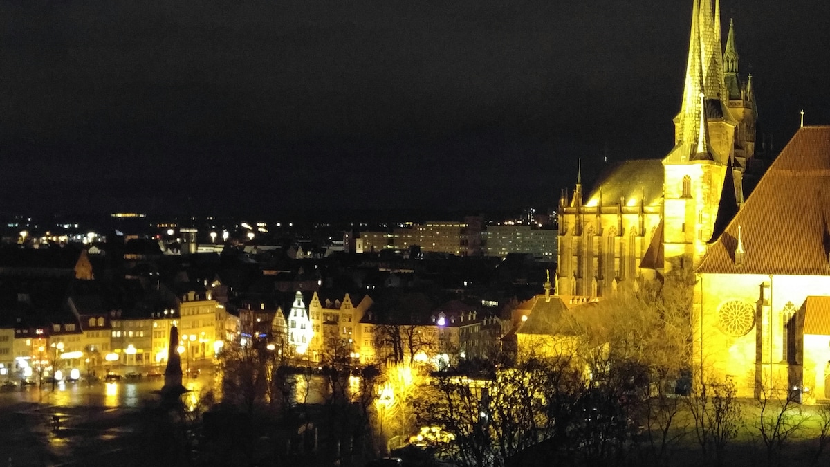 A night view reveals illuminated buildings in the distance, with warm lights reflecting off the surfaces. The prominent church features intricate architectural details, and trees are silhouetted against the night sky, enhancing the serene atmosphere of the cityscape.