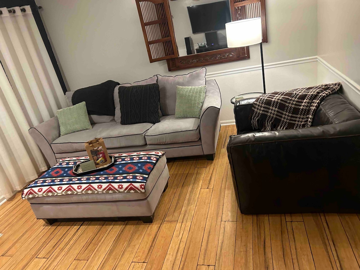 A cozy living room features a light gray sectional sofa adorned with green throw pillows and a patterned ottoman. A dark leather couch sits nearby. A decorative table holds a small centerpiece. Natural wood flooring complements the space, with a wall-mounted TV visible in the background.