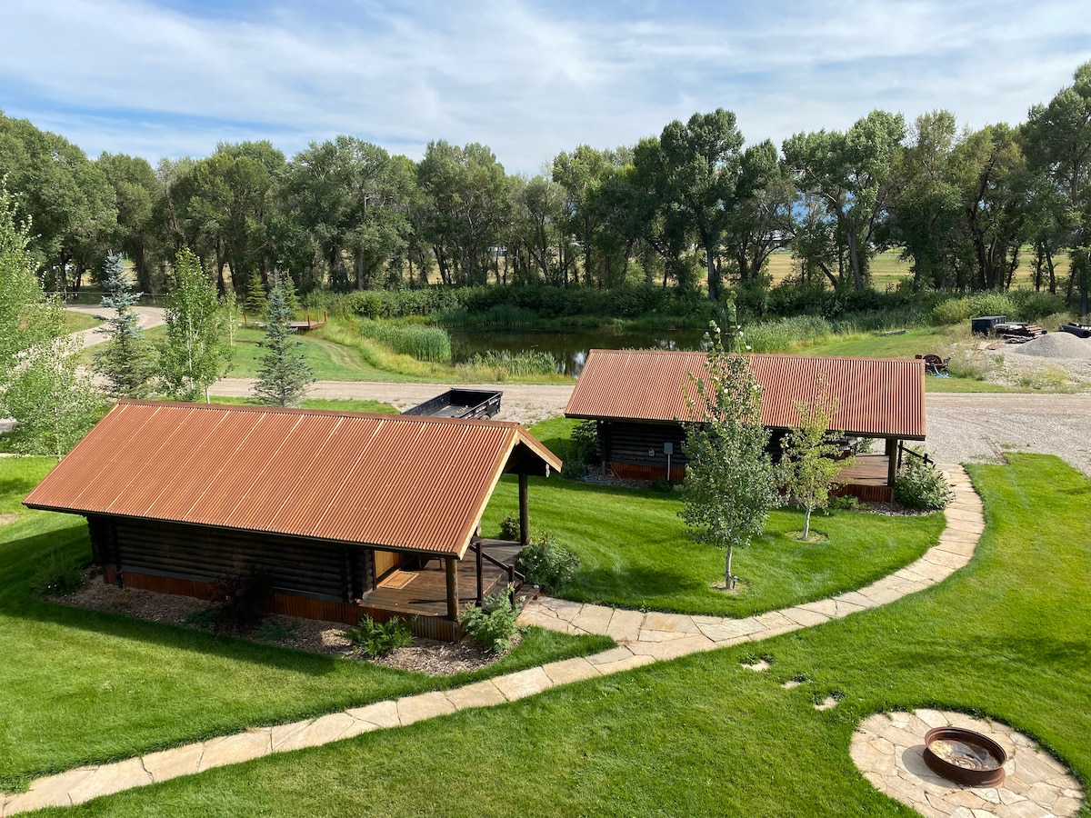 Two rustic cabins with orange metal roofs are set amidst a well-maintained lawn, surrounded by young trees. A curved stone pathway leads to the cabins, with grassy areas on either side. In the background, a stream and open fields are visible.
