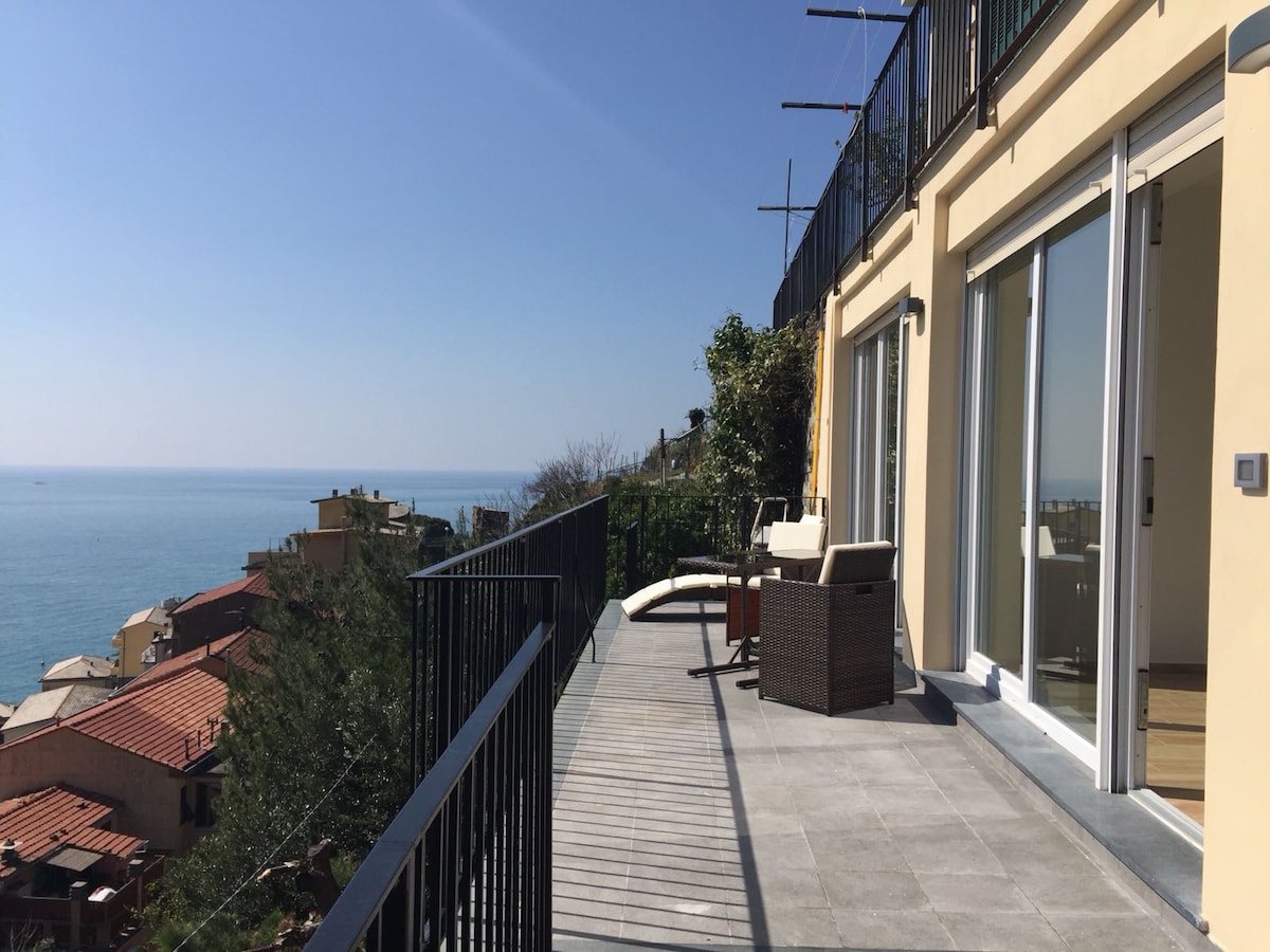 A spacious terrace features panoramic views of the sea, with contemporary outdoor furniture arranged for relaxation. The railing offers a clear view of the coastal landscape, while sliding glass doors lead into the interior living space.