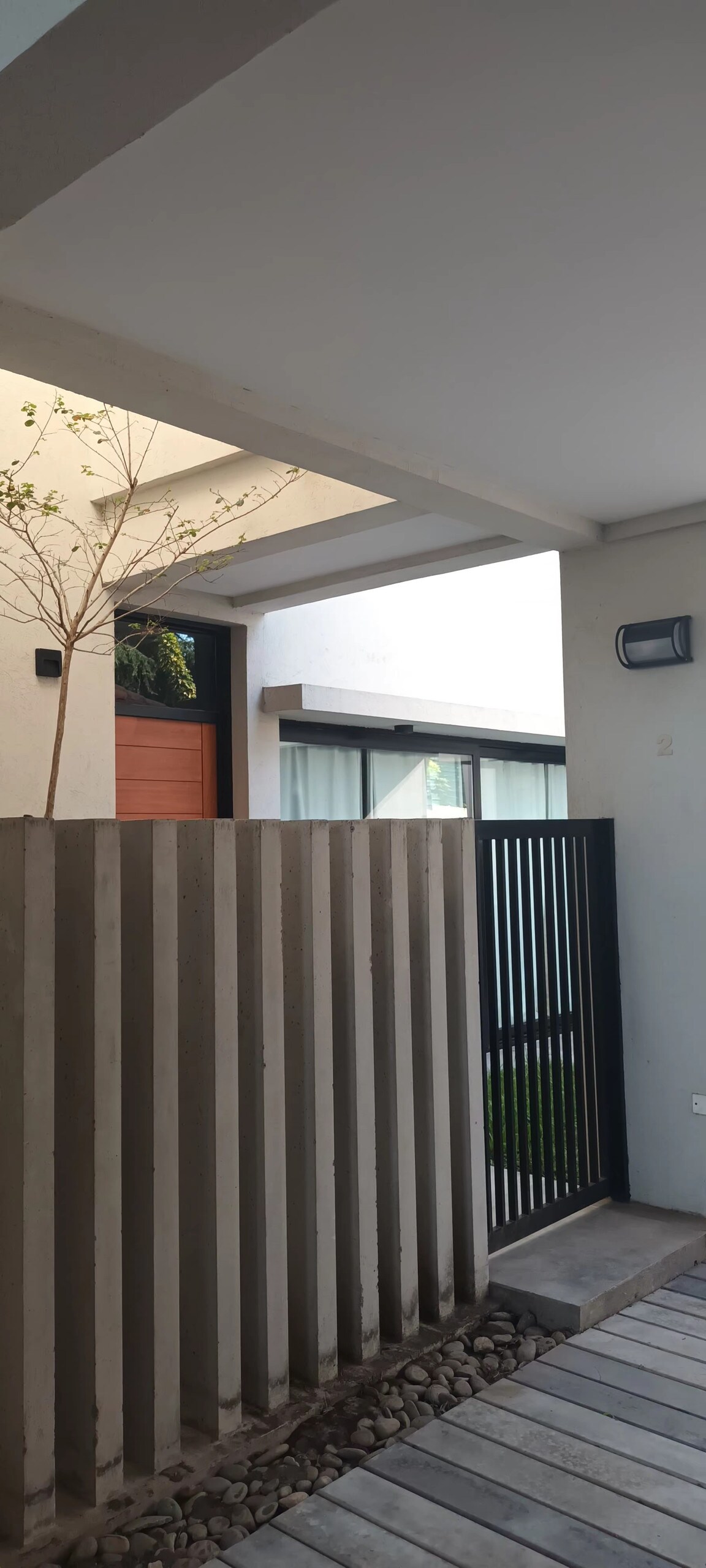 The entrance features a modern concrete fence with vertical lines. A small tree is situated beside the entrance, leading to a door framed by large glass panels. Soft natural light filters in, enhancing the welcoming ambiance of the space.