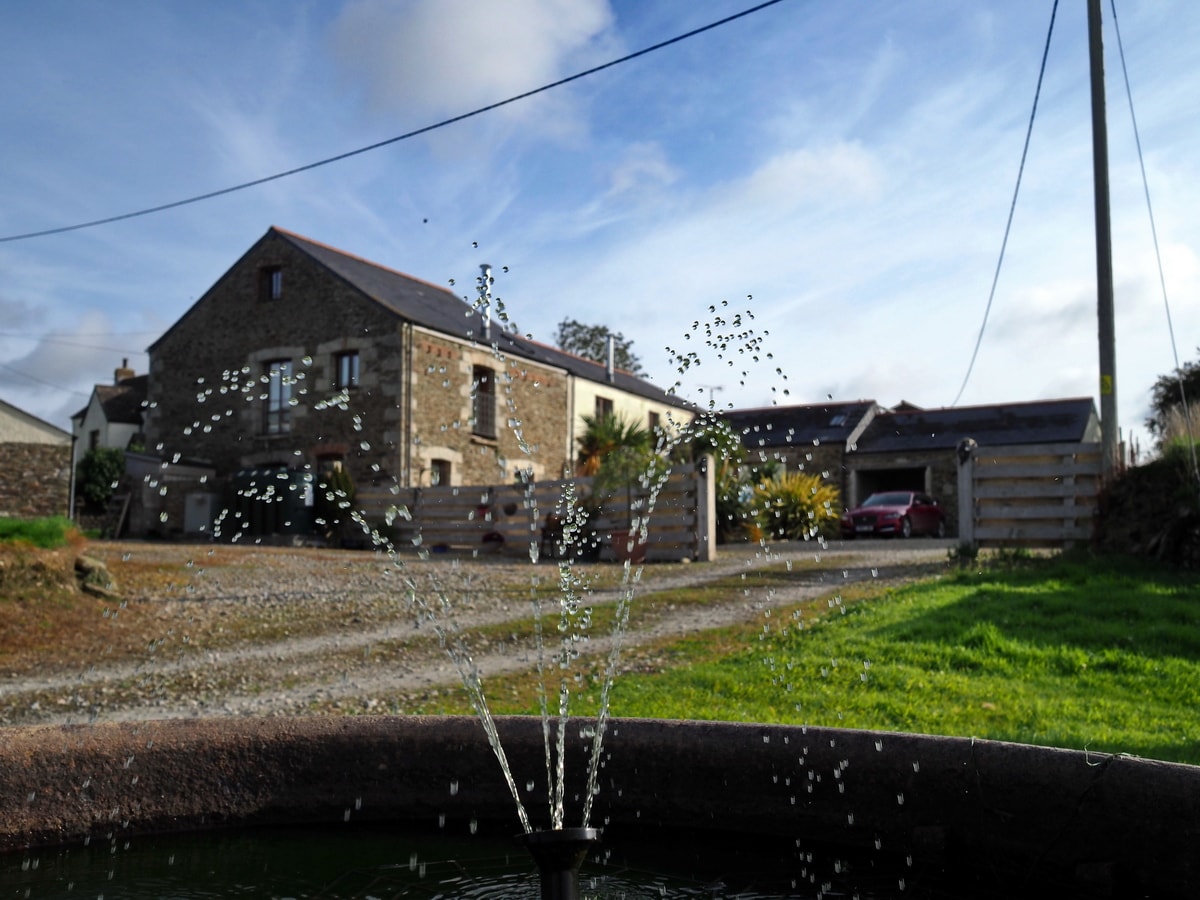 A stone barn conversion is featured in the background, surrounded by lush greenery and a gravel pathway. In the foreground, a fountain creates a gentle spray of water. A portion of the property with parked vehicles is visible to the right.