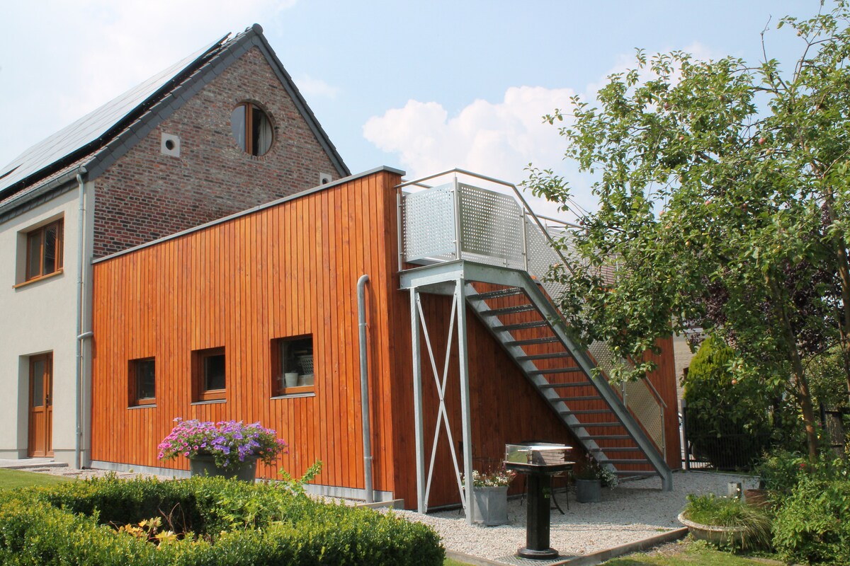 The exterior of a building combines wood and brick elements, featuring a staircase leading to a balcony. Flowering plants are situated near the base, and a garden area surrounds the structure, providing a serene setting.